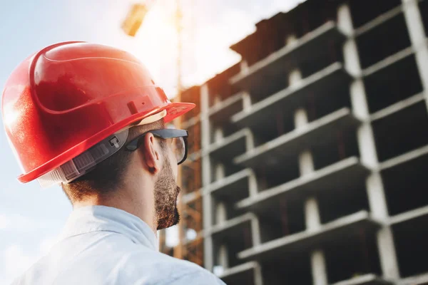 Young engineer in red helmet inspects construction of building. Bearded businessman on background of skyscraper under construction. Portrait of construction engineer on construction site background