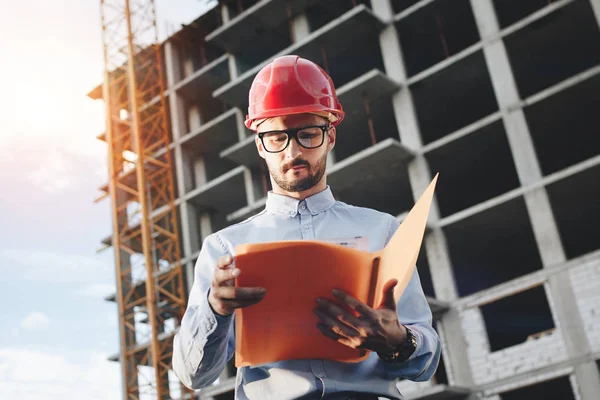 Joven ingeniero barbudo en casco de construcción rojo sobre el fondo de un edificio en construcción con dibujos y una carpeta en sus manos — Foto de Stock