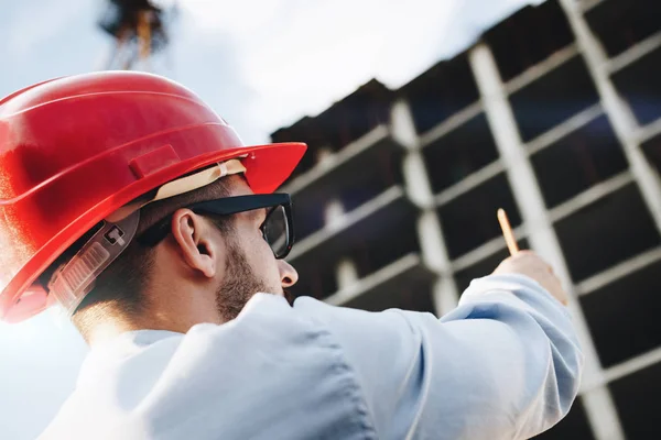 Unga skäggiga ingenjör eller arkitekt i röd hård hatt med penna i hand på byggarbetsplatsen. Verksamhet man hantera byggarbetsplats — Stockfoto