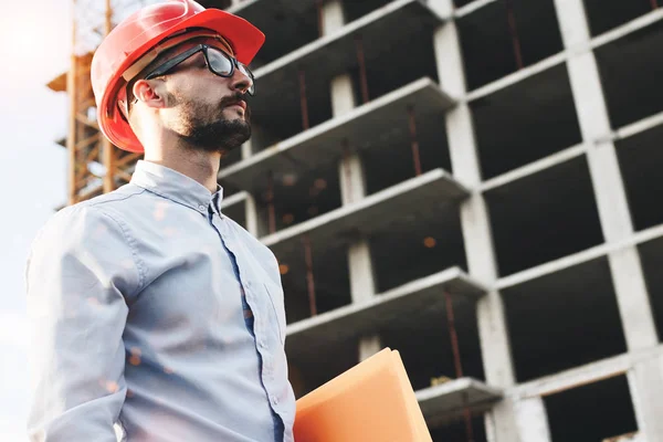 Giovane uomo d'affari barbuto o ingegnere che indossa occhiali e casco in cantiere. Ritratto di ispettore edile con cartella in mano sullo sfondo della costruzione dell'edificio — Foto Stock