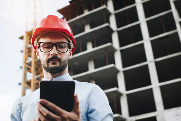 Engineer or architect in helmet with tablet in hand on construction site. Modern builder engineer in red hard hat with gadget in hand on building construction background — Zdjęcie stockowe