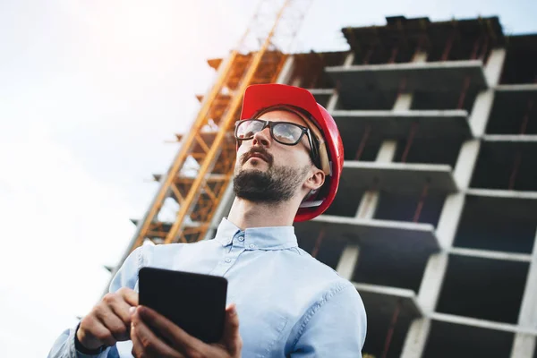 Joven ingeniero de casco rojo con tableta en las manos en el sitio de construcción. Inspector con tableta electrónica en obra —  Fotos de Stock