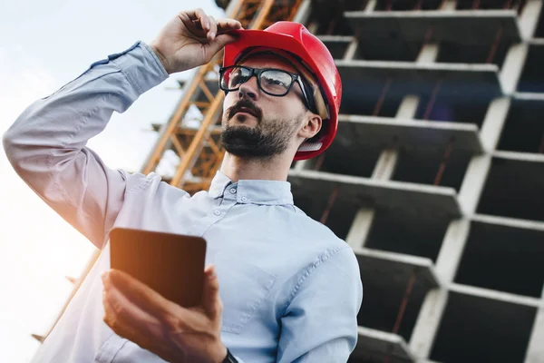 Barbudo joven ingeniero moderno constructor con aparato de tableta en la mano inspecciona la construcción de rascacielos. Concepto de ingeniero moderno e industria — Foto de Stock