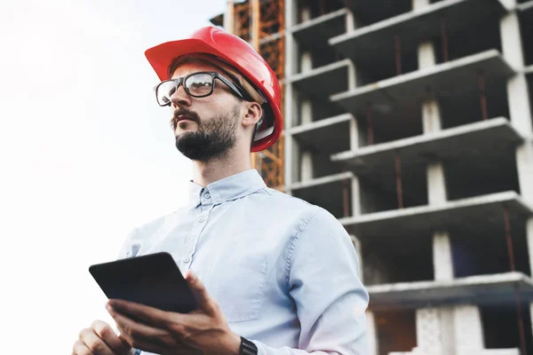 Businessman with glasses and hard hat with industrial tablet on the construction site. Industrial inspector with gadget on background of construction of plant — Stock Photo, Image