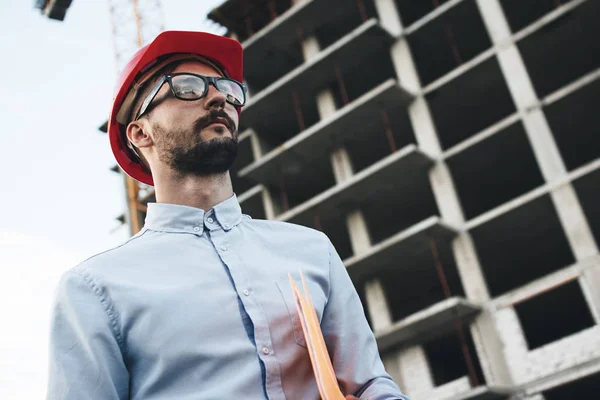 Modern business man in red hard hat holds folder with blueprint for construction industrial factory. Young bearded architect inspects construction of new building — Stock Photo, Image