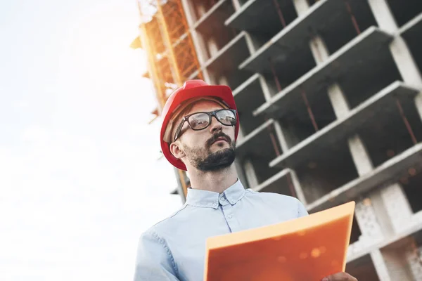 Ritratto di ingegnere maschio moderno in cantiere di nuovo impianto moderno. L'ispettore tiene una cartella con i documenti e controlla lo stato di avanzamento della costruzione dell'edificio. Lente flare effetc, sfondo sfocato — Foto Stock