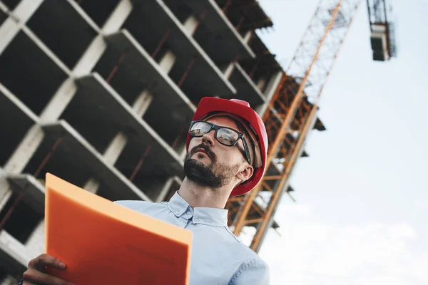 Giovane ingegnere moderno costruttore o architetto con cartella di documenti sullo sfondo dell'edificio in costruzione e gru. L'ispettore della sicurezza industriale controlla lo stato di avanzamento della costruzione dell'impresa industriale — Foto Stock