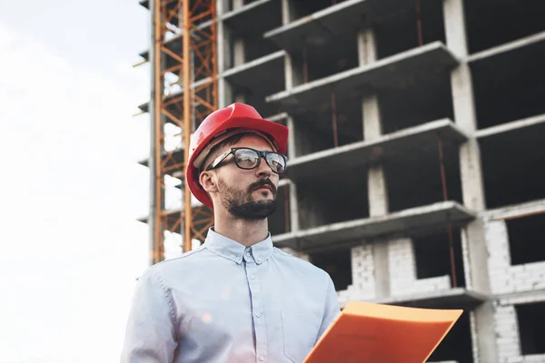 Il costruttore di ingegnere giovane e moderno tiene la cartella con disegni su sfondo di costruzione di edificio. Architetto in casco in cantiere — Foto Stock