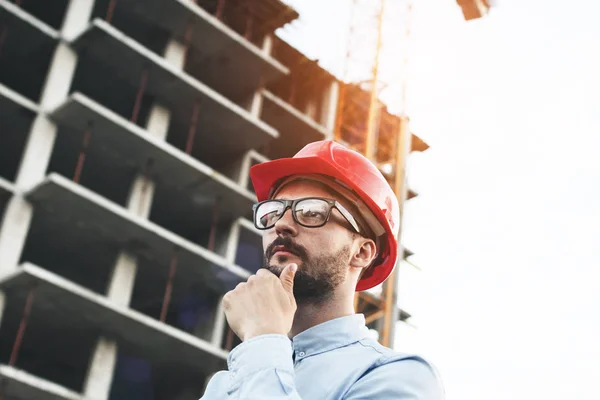 Thoughtful engineer in red hard hat thinks over construction building. Portrait of architect in glasses solving problems at construction site — Stock Photo, Image