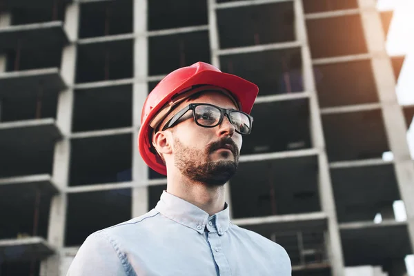 Joven constructor exitoso hombre de negocios con gafas y sombrero rojo duro en el sitio de construcción inspecciona la construcción de una nueva fábrica moderna — Foto de Stock