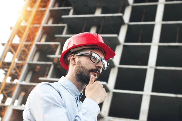 Ügyes építész vagy építési mérnök a consruction oldalon. Fiatal szakállas üzletember, háttérben a vasbeton csontváz felhőkarcoló — Stock Fotó