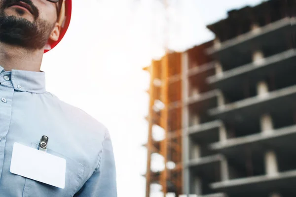 Blank white badge or business card on chest of a shirt of  modern engineer or architect in a red hard hat on background of  construction site. Young business man with blank white badge on chest of his shirt