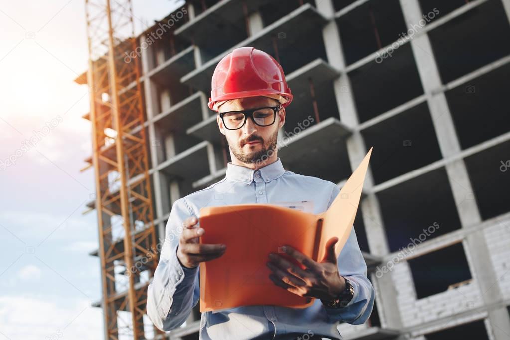 Young bearded engineer in red construction helmet on background of a building under construction with drawings and a folder in his hands