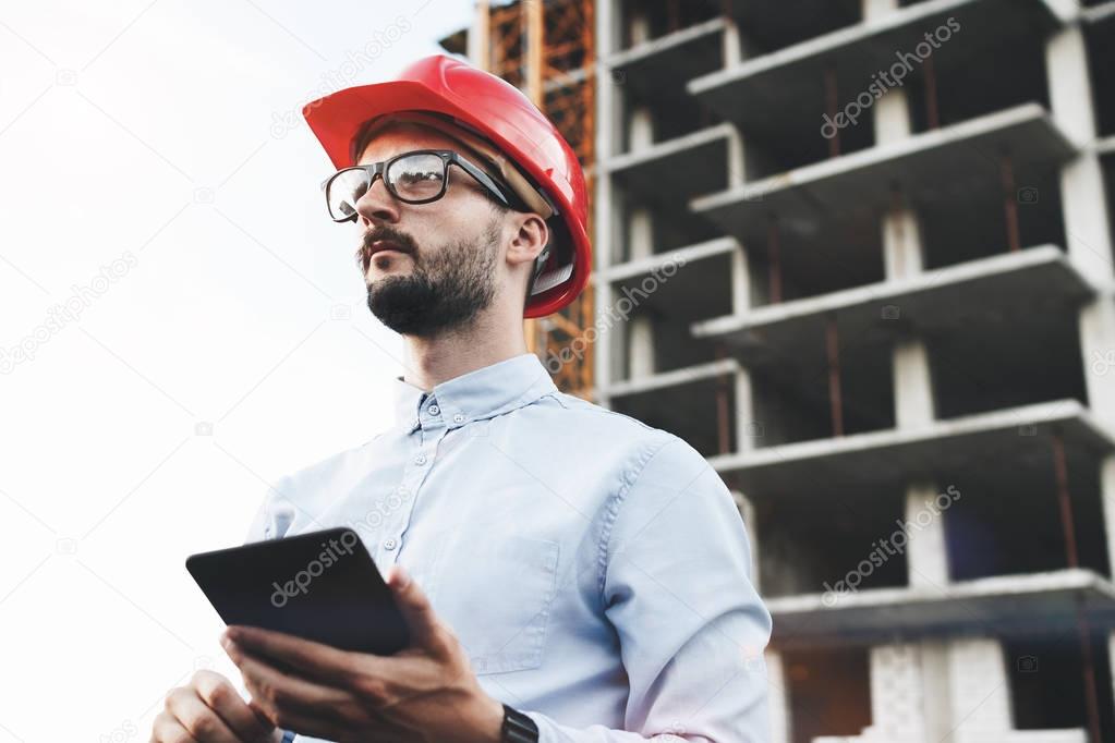 Businessman with glasses and hard hat with industrial tablet on the construction site. Industrial inspector with gadget on background of construction of plant
