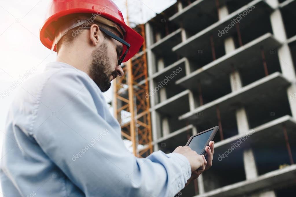 Bearded engineer in red hard hat with modern industrial electronic tablet on background of reinforced concrete skeleton of skyscraper. Close-up portrait of modern architect at work with gadget on construction site