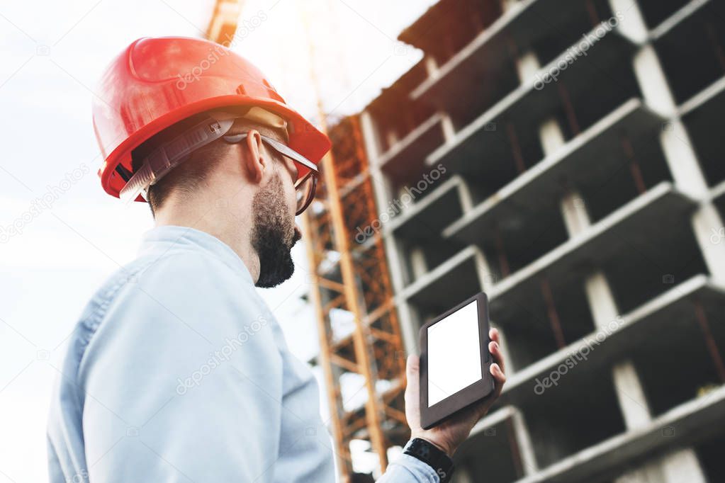 Engineer in helmet on construction of new modern enterprise with tablet in hand. Concept of modern architect or engineer of builder and constructor of industrial facilities
