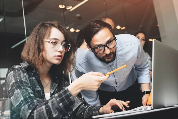 Ung och vacker affärskvinna i glasögon med penna i handen visar analys av projektet till sin skäggiga affärspartner. Unga medarbetare arbetar på ny start projekt i moderna kontor — Stockfoto