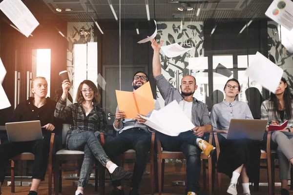 Gruppeglad hipster som jobber sammen og kaster opp papirdokumenter, grafer og har det gøy. Kreative coworking-personer med elektroniske innretninger og innretninger feirer vellykket gjennomføring av prosjektet – stockfoto