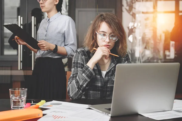 La arquitecta o diseñadora con gafas sostiene el lápiz en la mano y trabaja con el portátil moderno en el espacio del desván. Mujer de negocios que trabaja con documentos y dispositivos electrónicos. Bengalas intencionales de lentes — Foto de Stock