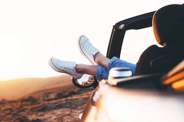 Modern hipster girl resting in convertible and admiring view. Woman driver watching sunset in cabriolet put feet on car door — Stock Photo, Image