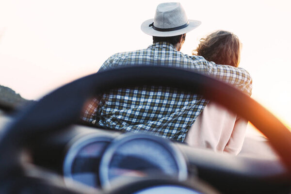 Trendy love couple hipsters sit on hood of cabriolet car and enjoy sunset in mountains