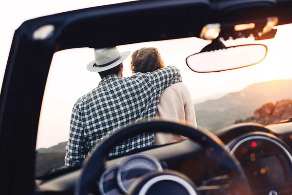 Pareja en el amor abrazos y se sienta en la capucha de cabriolet. Los viajeros masculinos y femeninos descansan y disfrutan del atardecer cerca de su coche — Foto de Stock