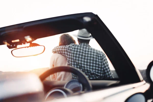 El hombre con sombrero y novia están descansando y mirando cerca de cabriolet al atardecer en las montañas. Pareja cariñosa de viajeros en coche disfrutar de la vista y se sienta en la capucha del descapotable — Foto de Stock