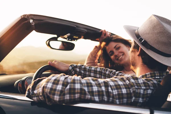 Loving couple of hipsters in cabriolet car enjoy view at sunset. Man in hat and passenger woman look at each other in convertible — Stock Photo, Image