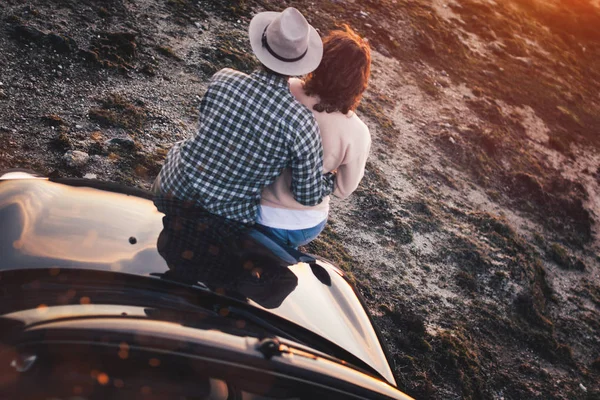 Loving couple sits on hood convertible car and enjoy view at sunset. Young trendy hipsters near cabrio look at sunset. Lens flare effect — Stock Photo, Image