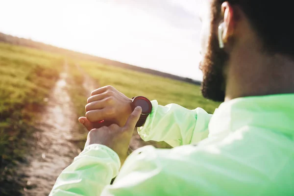 Atleta con auriculares bluetooth mira reloj inteligente y analiza los resultados del entrenamiento al aire libre. Concepto deportista moderno con gadgets — Foto de Stock