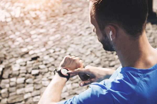 Joven atleta barbudo con auriculares bluetooth analiza el resultado del entrenamiento en smartwatch. Concepto de atleta moderno con reloj inteligente y gadgets en el entorno urbano — Foto de Stock