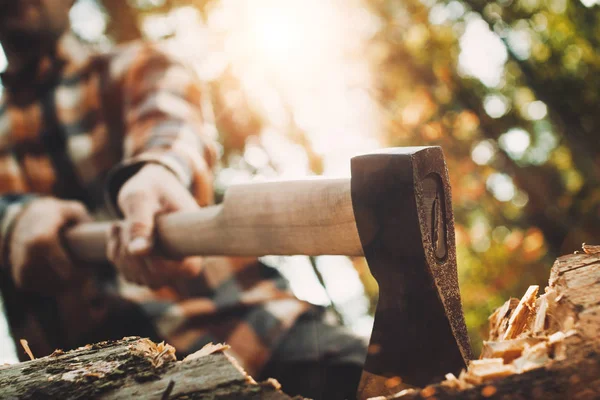 Fuerte leñador con camisa a cuadros sosteniendo hacha en sus manos y cortando árboles en el bosque. Axe de cerca. Fondo borroso, efecto de destello de lente — Foto de Stock