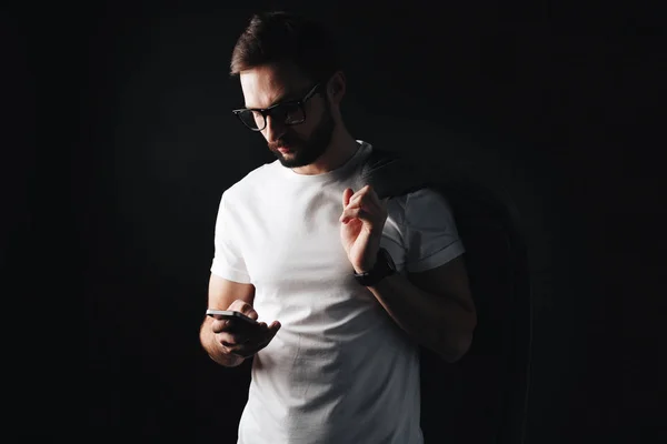 Hombre barbudo joven en gafas y abrigo se ve en el teléfono inteligente en camiseta blanca. Moda hipster con camiseta blanca en blanco sobre fondo negro — Foto de Stock