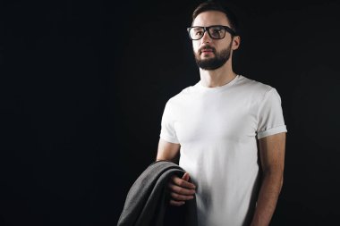 Sakallı yakışıklı bir beyaz boş tshirt poz ve onun el arka plan siyah duvar üzerine bir kat holding gözlük
