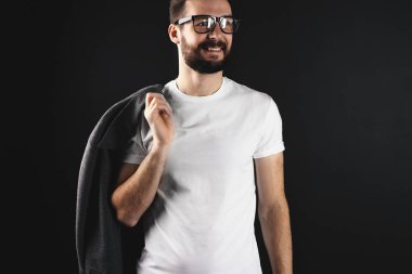 Koyu arka plan üzerinde beyaz boş t-shirt giyen omuz montlu yakışıklı hipster gülümseyerek. Sakallı adam gözlük ve boş tshirt 