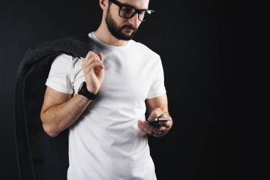 Gözlük ve ceket omzunun beyaz boş t-shirt giyen üzerinde ciddi sakallı işadamı smartphone vasıl görünüyor