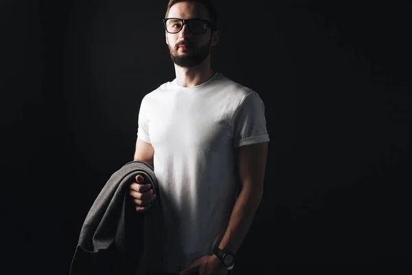 En tom vit t-shirt på unga skäggiga affärsman stående på en svart bakgrund. Hipster med en kappa och glasögon poserar i en vit tshirt — Stockfoto