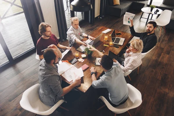 Grupo de personas coworking sentados alrededor de la mesa de madera y trabajando en las computadoras portátiles modernas en el loft. Los gerentes analizan documentos comerciales y utilizan tabletas táctiles. Concepto de reunión de negocios — Foto de Stock