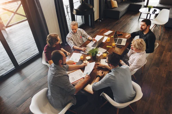 Employees of creative company are working on new startup and discussing idea of business development. Coworkers sit around wooden table and work on laptops and tablets. Concept of work in loft space — Stock Photo, Image