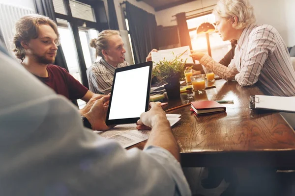 Uomo che tiene tablet con schermo bianco in mano e discutere con il team di coworking nuovo progetto creativo. Uomini d'affari seduti intorno al tavolo nello spazio soppalco — Foto Stock