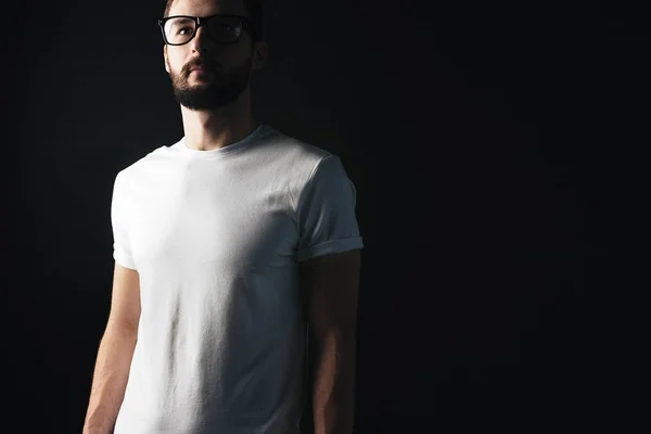 Młody Brodaty hipster w okularach, ubrany w biały t-shirt puste z cień na jego twarz na ciemnym tle — Zdjęcie stockowe