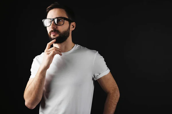 Joven barbudo pensativo hombre con gafas vestidas con una camiseta blanca sobre un fondo negro —  Fotos de Stock