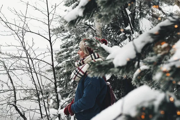 Довольно счастливая женщина хипстер в красной шляпе и клетчатый шарф стоя в диком лесу перед удивительным видом на зиму — стоковое фото