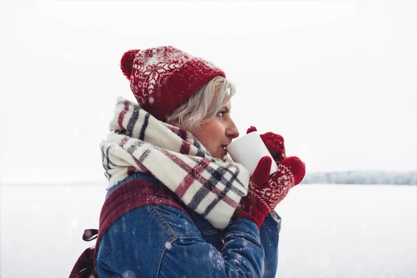 Красивая хипстерша в зимней одежде с чашкой горячего кофе на фоне снежного пейзажа — стоковое фото