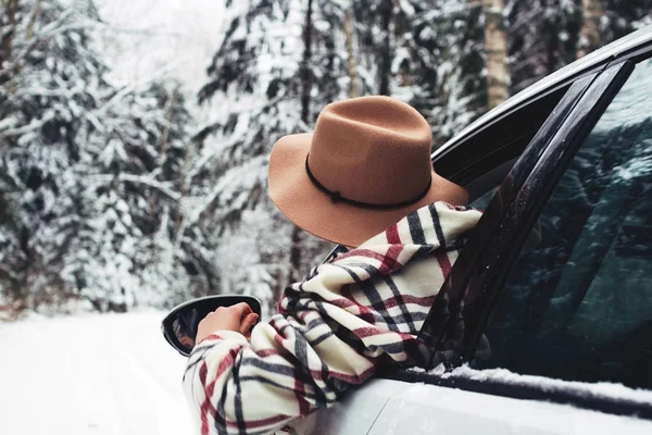 Stilig resande kvinnan njuter av vintern väg och snö skogen från bilfönstret. Unga hipster flicka klädd i mössa och halsduk — Stockfoto