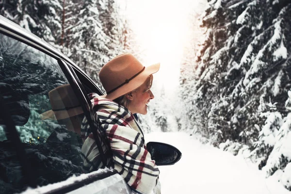 Lycklig kvinna resenären njuter en snöig bild av en skog vinterväg från bilfönstret. Unga hipster flicka i vintage hatt hängande ur bilfönstret — Stockfoto