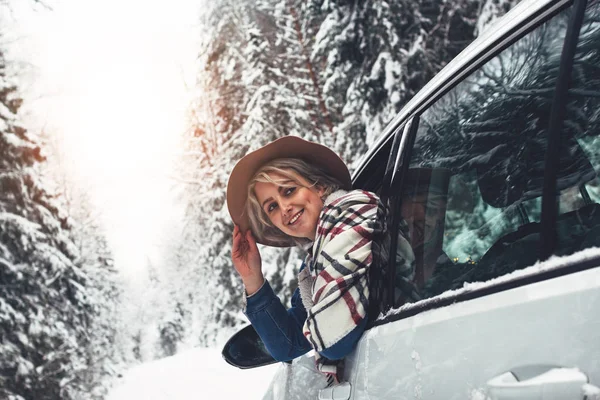Vacker och söt hipster tjej bär hatt och rutig scarf i bil njuter av vintern skogsväg. Kvinna som reser i Sportlov på bil — Stockfoto