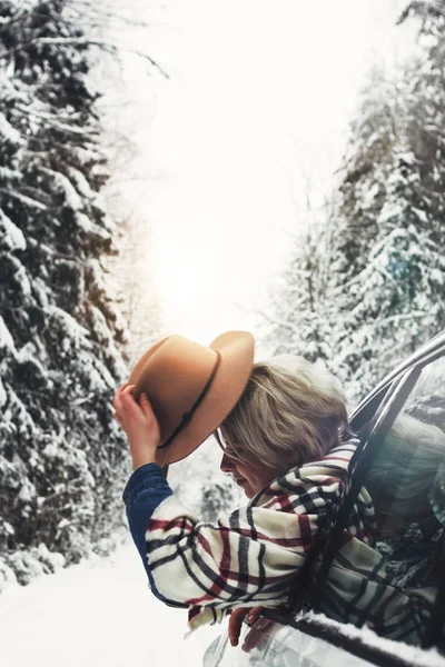Vacker hipster kvinna i hatt och rutig halsduk hängande ur bilfönstret. Söt flicka reser på vinterväg i snö skog — Stockfoto