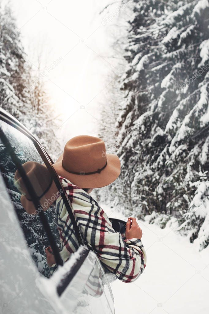 Handsome woman traveler sits in the car and enjoying a beautiful winter view of the forest road. Wearing hat and checkered scarf. Winter holidays. Wanderlust