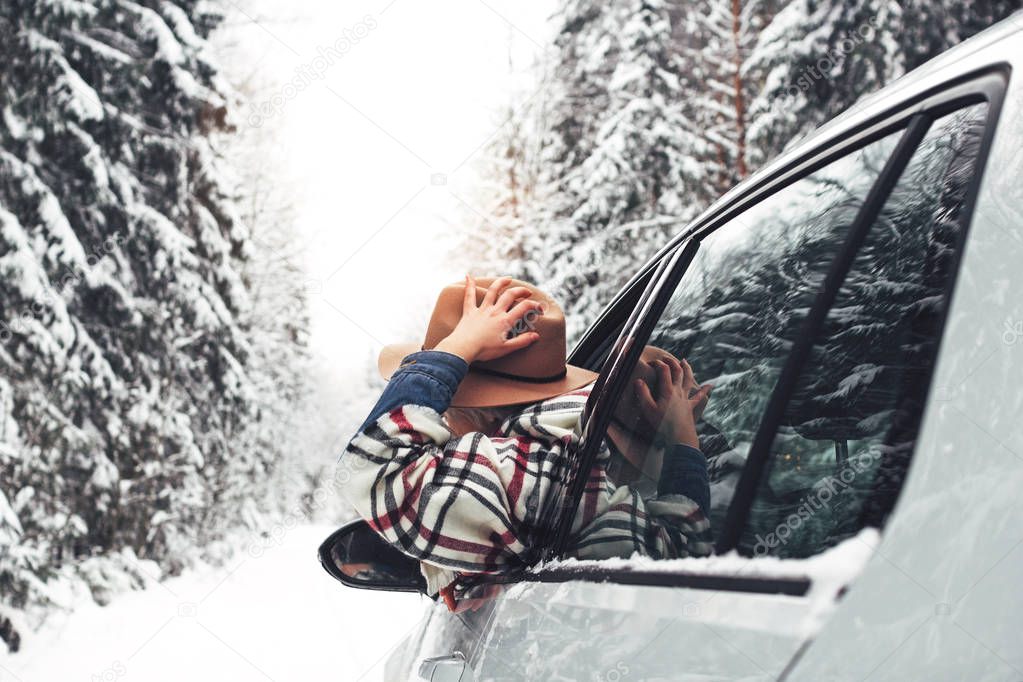 Handsome woman hanging out of the car window, hold vintage hat and enjoying beautiful winter snow road in forest. Traveler in winter nature